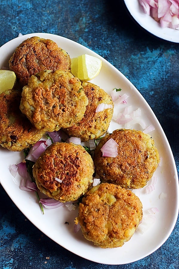 Crispy street style aloo tikki served in a ceramic plate topped with finely chopped raw onions and lemon wedges