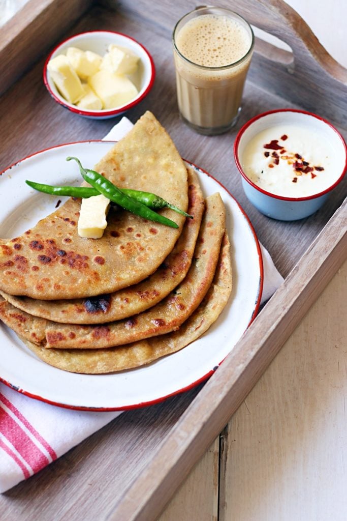 Fresh homemade aloo paratha stcaked with some butter and served with chai, dahi and more butter on a wooden tray