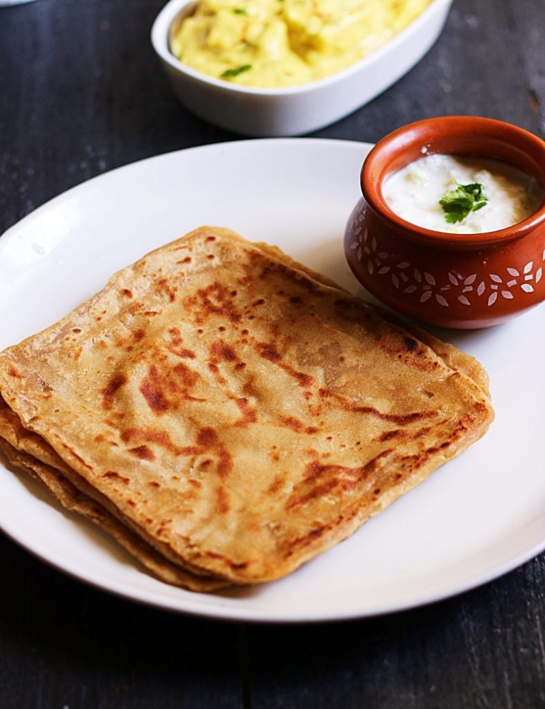 soft flaky parathas stacked in a white plate and served with yogurt and shahi paneer