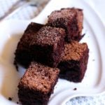 small squares of eggless chocolate cake dusted with sugar served on a white snack plate.