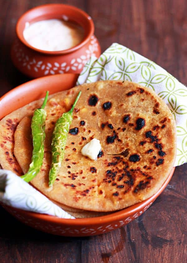 wholesome and delicious Punjabi Paneer Paratha served on brown plate with fried chilies and a dab of butter. Some thick yogurt in a small bowl is served as side dish.