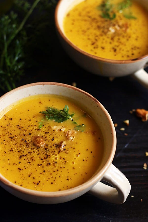 Closeup shot of vegan pumpkin soup served with toasted walnuts garnish in white soup bowl. In the background there is one more serving of pumpkin soup