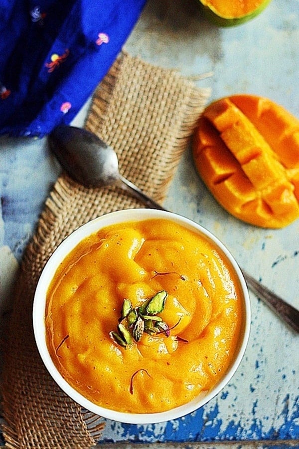 Homemade aamras served in a small white ceramic bowl with a spoon. Ripe mangoes in the background.