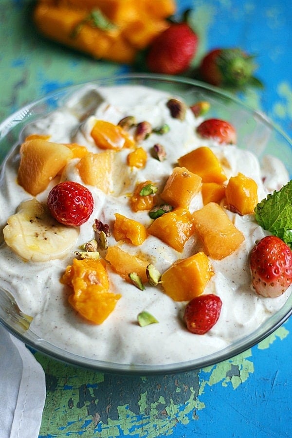 A glass bowl full of chilled fruit cream- an Indian fruit dessert with more fruits in background.