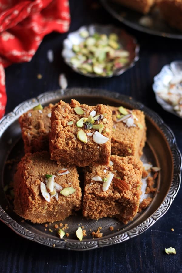 mohanthal mithai served in a silver plate. There are chopped nuts placed in small pinch bowls in the background