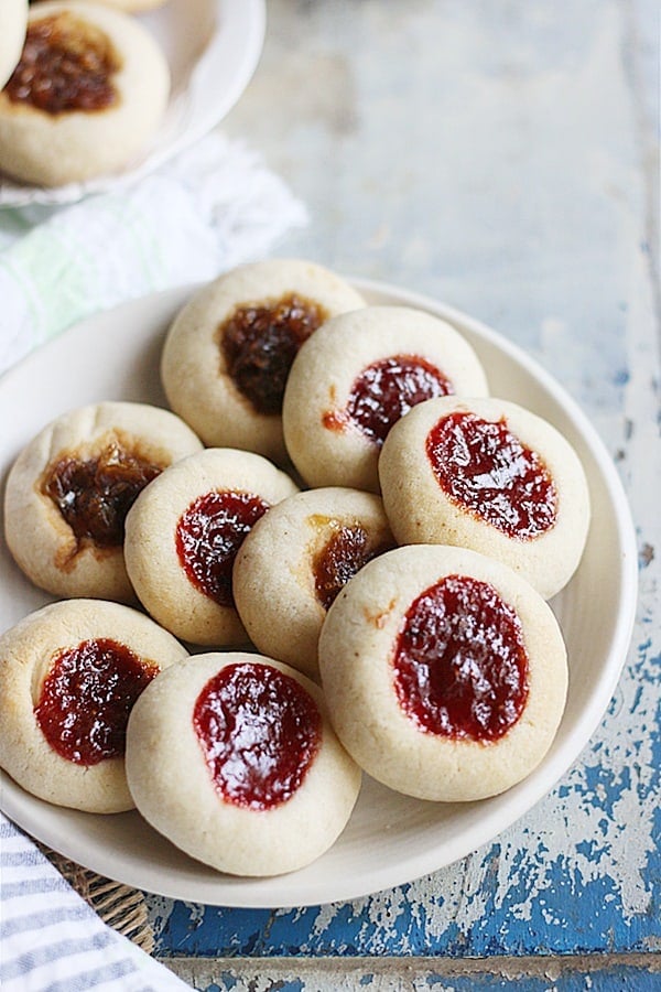 Homemade thumbprint cookies arranged in a white ceramic plate with more cookies in background.